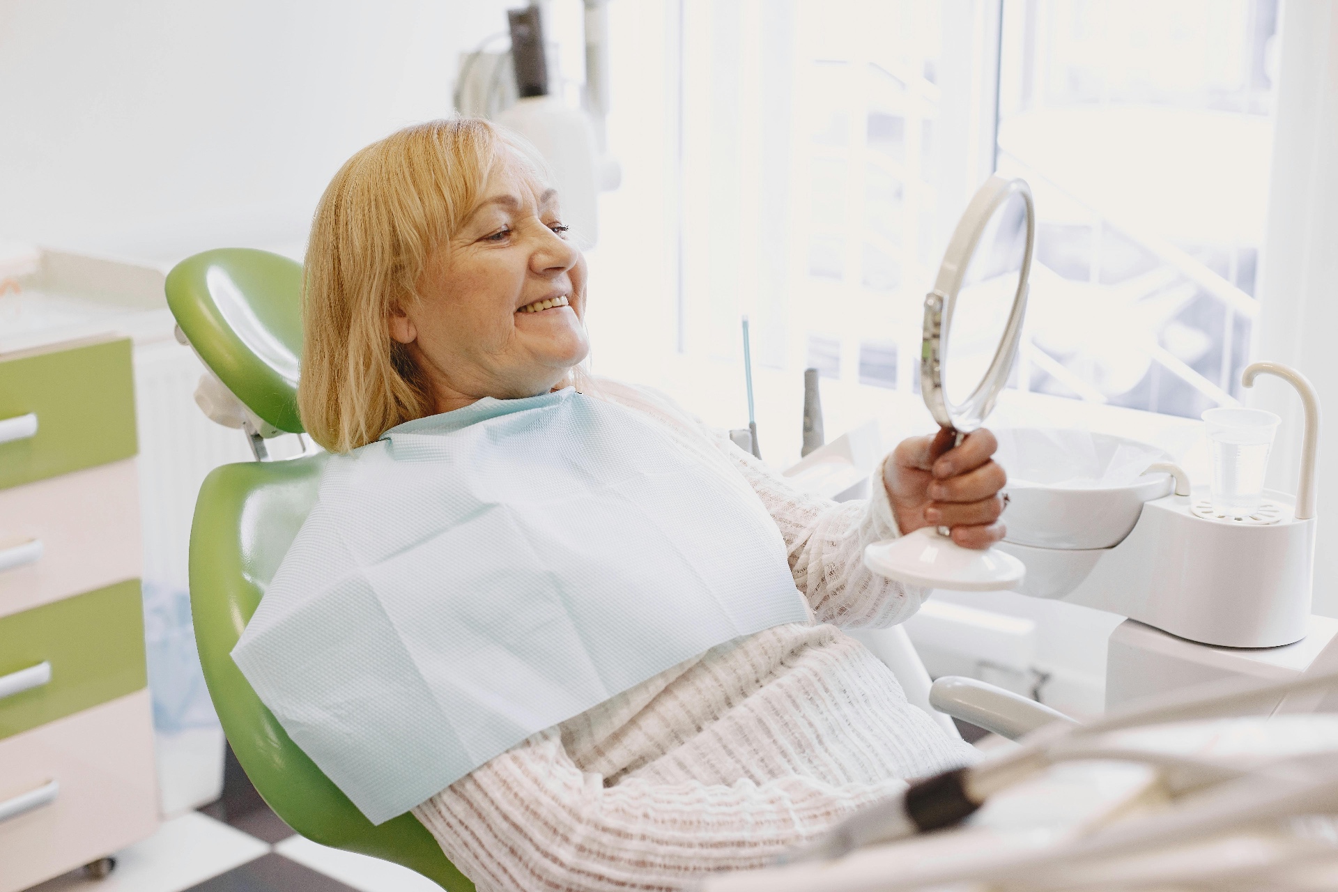 An older dental patient smiling while looking in a hand mirror in the treatment chair